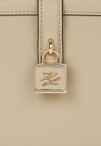 Beige színű táska sima textúrával, arany zárral és emblémával. A dizájnja strukturált vonalakkal és kontrasztos varrással rendelkezik.