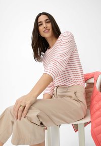 Gestreiftes Langarmhemd in Weiß mit roten Streifen, kombiniert mit beigen weiten Hosen. Eine gesteppte korallenfarbene Jacke liegt über einem Stuhl.