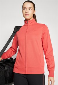 Koralinė zip-up sportinė striukė, pagaminta iš minkštos medžiagos, su ilgomis rankovėmis, aukštu apykakle ir logotipu krūtinėje. Juoda sportinė kuprinė rankoje.