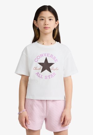 Jeune personne portant un t-shirt blanc Converse All Star et un short rose clair, debout avec une expression neutre sur un fond uni.