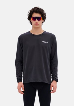 Mann trägt schwarzes Langarmshirt mit „cep“-Logo, schwarze Leggings, bunte Sportsonnenbrille mit Rundumrahmen und eine schwarze Armbanduhr, steht frontal.