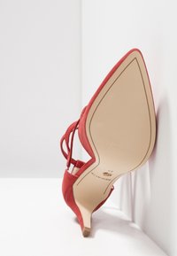 Richelieu rouge en velours avec un talon haut, à bout pointu et brides de cheville. La semelle est beige avec un marquage doré et une texture lisse.