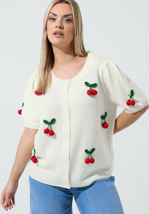 Femme portant un cardigan blanc boutonné avec des motifs de cerises rouges et un jean bleu, debout devant un fond clair uni.