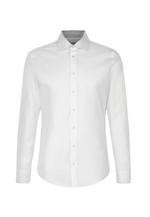 Homme portant une chemise blanche à boutons, un pantalon ajusté foncé, des chaussures habillées noires, debout les mains dans les poches devant un fond uni.