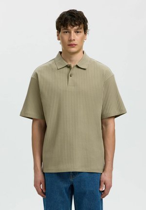 Tânăr purtând un tricou polo bej cu mâneci scurte și dungi și blugi albaștri, stând în fața unui fundal simplu, deschis.