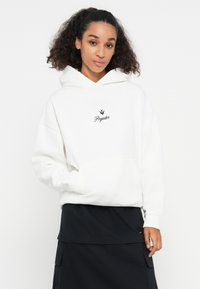 Hoodie en coton blanc avec une poche frontale et un logo brodé noir "Pegador". Associé à une jupe noire à plusieurs couches.