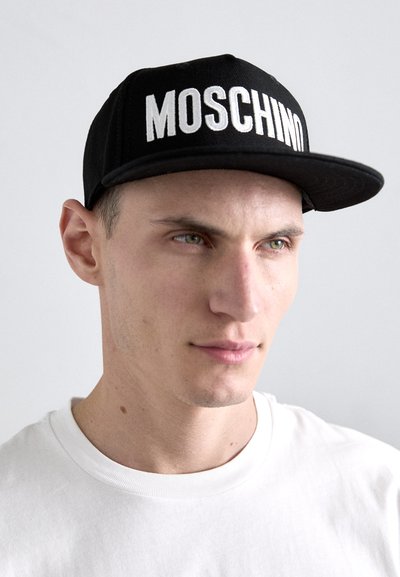 Casquette de baseball noire avec "MOSCHINO" en grandes lettres blanches; portée avec un t-shirt blanc à col rond.