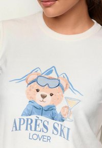 Bjørnebamse iført blå skibriller og jakke, der holder et cocktailglas med bjerglandskab i baggrunden og teksten "APRÈS SKI LOVER".