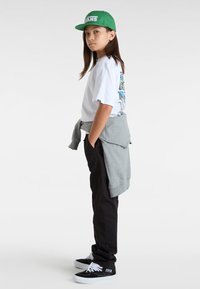 Vit t-shirt med grafisk tryck, grå sweatshirt knuten runt midjan, svarta byxor och svarta skor med vita skosnören; grön keps med logotyp.