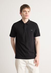 Sort bomulds polo skjorte med en to-knap halsudskæring og korte ærmer. Har et lille mærke på venstre bryst. Enkel design, glat tekstur.