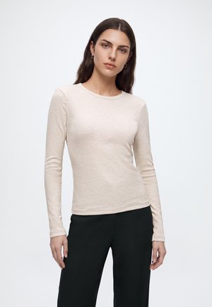 Femme aux longs cheveux foncés portant une chemise beige ajustée à manches longues et un pantalon noir, debout devant un fond blanc uni.