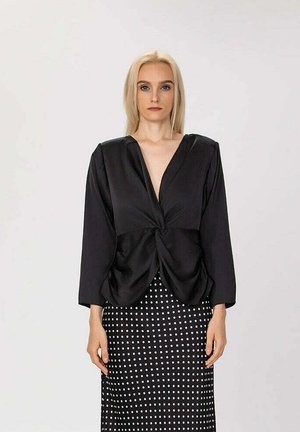 Femme blonde portant un chemisier noir torsadé à l'avant et une jupe noire à pois blancs, debout devant un fond clair uni.
