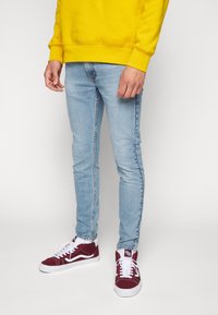 Vaqueros pitillo azul claro combinados con zapatillas burdeos y una sudadera de color amarillo brillante, con un escote redondeado y mangas largas.