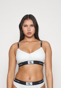 Bijela grudnjak od glatke tkanine, s potporom od žice i crnim Calvin Klein logom na trakama i pojasu. Jednostavan, minimalni dizajn.