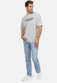 Šedé tričko s krátkým rukávem s grafikou "TOP GUN" v červené a modré barvě, v kombinaci s světle modrými džínami a bílými teniskami. Řídké pozadí.