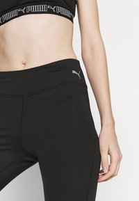 Črne športne legice z visokim pasom, ki imajo sivo Puma logo na levem bok, gladko tkanino in eleganten dizajn.