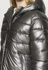 Metalická šedá prošívaná bunda s kapucí, s prošívanými částmi, zapínáním na zip a bočními kapsami. Hladký, lesklý povrch a nastavitelný detail.