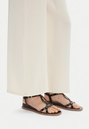 Schwarze Ledersandalen mit T-Spangen-Design, dekorativen Nieten und flacher Sohle, getragen zu hellfarbenen Hosen mit weitem Bein.