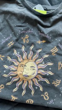Mørkegrå T-shirt med et centralt solmotiv med lilla og guld accenter, stjernetegnssymboler og detaljerede ansigtstræk integreret i solen.