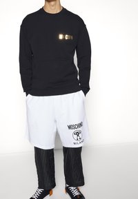 Černá mikina s logo "MOSCHINO" ve zlaté barvě, v kombinaci s bílými šortkami s černým logem a proužkovanými kalhotami. Černé sportovní boty s oranžovými akcenty.