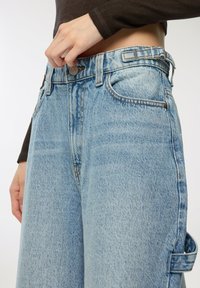 Jeans a gamba larga in denim azzurro chiaro con vita alta, dotati di chiusura a bottone, passanti per cintura e tasche laterali. Tessuto testurizzato con un aspetto sbiadito.
