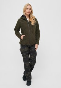 Olivengroene fleecejas met een capuchon en ritssluiting, gecombineerd met zwarte camouflage cargo-broeken met meerdere zakken en een relaxte pasvorm.