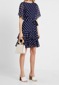 Marinblå prickig klänning med utsvängd nederkant och knytmidja. Har genomskinliga ärmar och lager av volanger. Kompletterad med en beige handväska och vita sandaler.