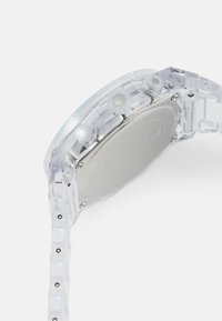 Permatomas plastikinis laikrodis su apvaliu cichlu, sidabriniu metalu ir mygtukais. Dirželis turi segmentuotą dizainą, rodantį lygų paviršių.