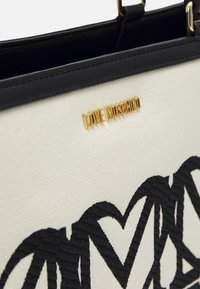 Vitt kanvas handväska med svart broderad hjärtpatron, med en guld "LOVE MOSCHINO"-logotyp och svarta läderdetaljer.