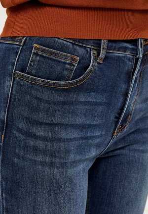 Tumedat sinised teksad klassikalise lõikega, millel on oranž õmblus, väike eesmine tasku ja nupuga sulgur. Sile tekstuur.