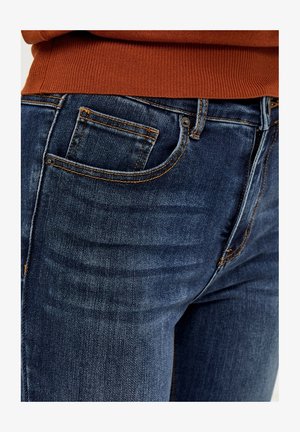 Tumedat sinised teksad klassikalise lõikega, millel on oranž õmblus, väike eesmine tasku ja nupuga sulgur. Sile tekstuur.