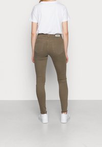 Jeans skinny verde oliva con una cintura alta, bolsillos traseros y una etiqueta etiquetada "ONLY", combinados con una camiseta blanca y zapatillas blancas.