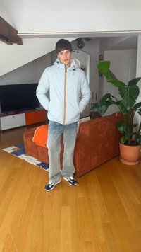 Jonge man in een lichtblauw jasje en grijze spijkerbroek staat in de woonkamer met houten vloer, oranje bank, grote potplant en tv op de achtergrond.