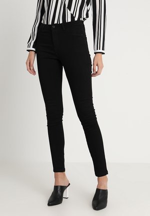 Femme portant un pantalon skinny noir, des mules noires à talons blocs blancs, et un haut à manches longues rayé vertical noir et blanc.
