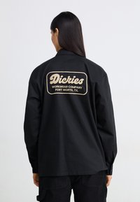 Maglietta a maniche lunghe nera realizzata in un mix di cotone, con un logo ricamato beige sulla schiena. Presenta un colletto e polsini con bottoni.