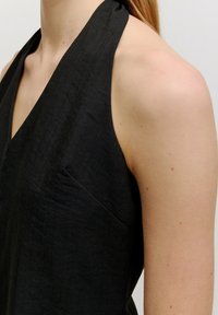 Robe noire à col licou avec un décolleté plongeant en V, en tissu texturé et un détail subtil avec une poche latérale. Teint de peau lisse visible.