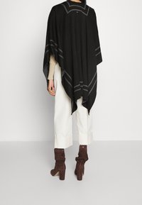 Svart poncho med grå randdetaljer, har en lös design och är gjord av mjukt tyg. Bärs över cremefärgade byxor och bruna slappa stövlar.