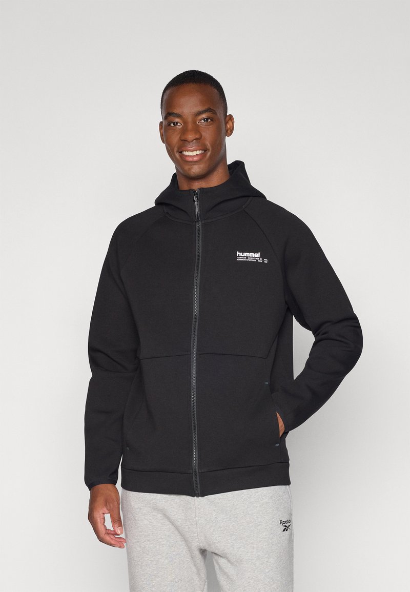 Sweat à capuche noir zippé en tissu doux, doté d'une capuche, de manches raglan et d'un petit logo blanc sur la poitrine gauche. Porté avec un pantalon de jogging gris.