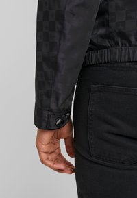 Svart kviltad jacka med rutmönster, tryckknappsstängning vid manschetten och elastisk nederkant, kombinerad med svarta byxor.