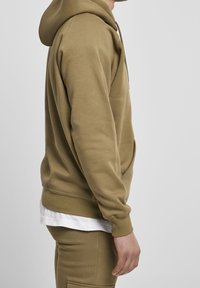 Olivově zelená mikina s kapucí z bavlněné směsi, vybavená stahovací kapucí, přední kapsou a žebrovanými lemy na rukávech a spodním okraji. Nositelná přes bílou košili.