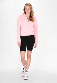 Lichtroze cropped zip hoodie met voorkruimelzak, gecombineerd met zwarte fietsshorts en witte sneakers met zwarte accenten.
