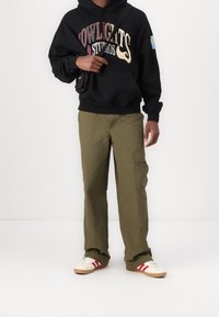 Schwarzer Hoodie mit der Aufschrift "NOWLIGHTS STUDIOS", olivfarbene Cargohose mit Seitentaschen und weiße Sneakers mit roten Streifen.