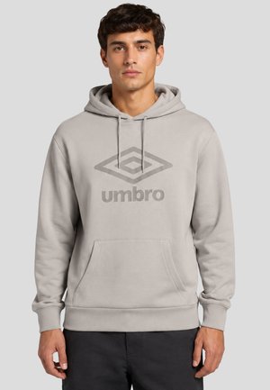 Giovane uomo con indosso una felpa con cappuccio Umbro grigio chiaro con tasca frontale e cappuccio con coulisse, in piedi davanti a uno sfondo grigio uniforme.
