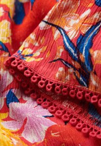 Tessuto floreale con uno sfondo rosso vivace e fiori gialli e blu. Decorato con un bordo in pizzo testurizzato a loops lungo il margine.