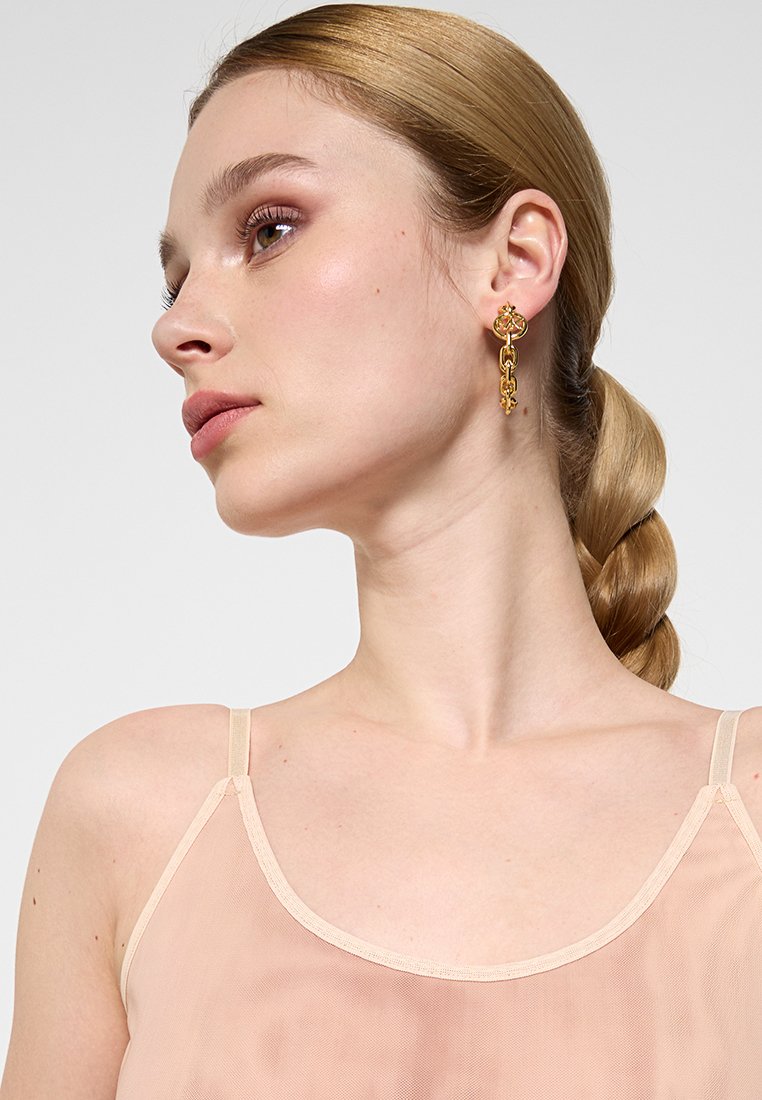 Gouden oorbellen met bloemaccenten en een geschakelde kettingontwerp, gedragen met een doorschijnende perzikkleurige top met dunne bandjes. Glad oppervlak en glanzende afwerking.