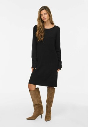 Femme aux longs cheveux portant une robe noire en maille à manches longues et des bottes marron en daim à talons montant jusqu'aux genoux, debout sur un fond blanc.