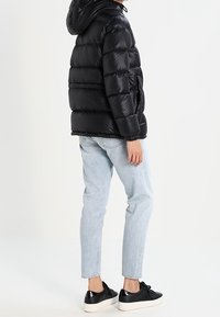 Svart vadderad jacka med huva och kviltad design, bärs över ljusblå jeans och svarta sneakers, och visar en slät, glänsande yta.