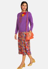 Frau trägt einen lila Pullover, einen floral gemusterten Midirock, goldene Schuhe, eine orangefarbene Handtasche und eine Kette mit roten und orangenen Blumen vor einem weißen Hintergrund.