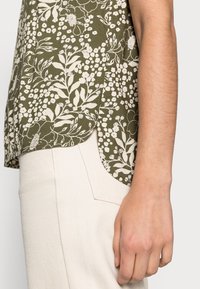 Vista lateral de una persona que lleva una camisa floral verde olivo y pantalones beige claros, con el brazo derecho colgando relajado.