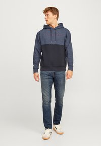 Sudadera azul marino con una sección superior de un azul más claro y texto rojo. Presenta una capucha con cordón, puños acanalados y una textura suave. Se lleva con vaqueros y zapatillas.